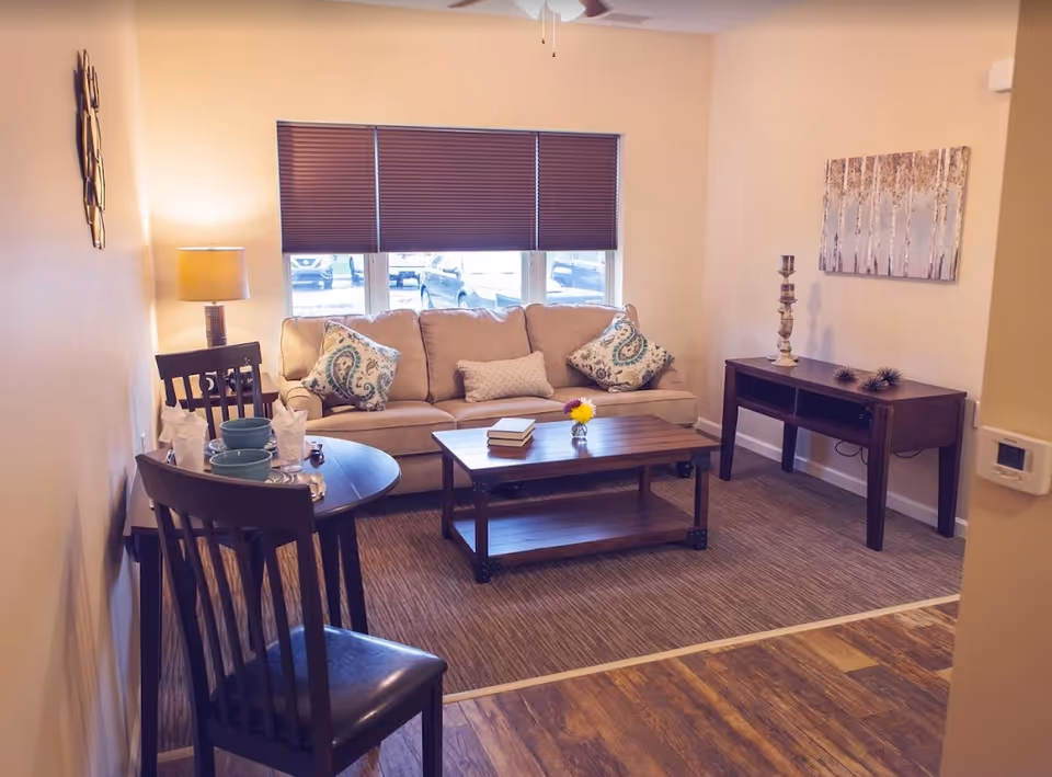 A cozy living room with a beige sofa adorned with patterned cushions, a wooden coffee table with books and a small flower vase, a side table with a lamp, and a small dining table set with plates and napkins. The room has a window with closed blinds and light-colored walls with a piece of artwork and a decorative candle holder on a console table.