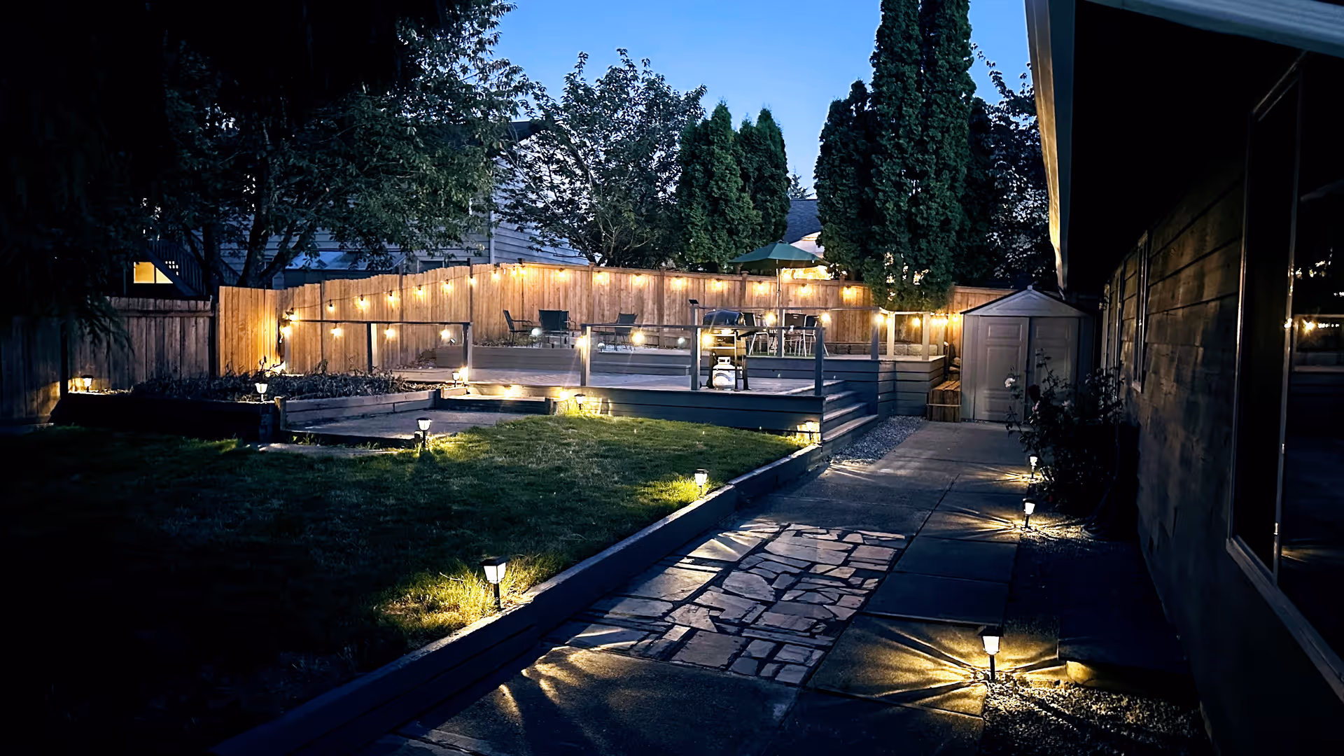 A backyard at dusk with a well-lit pathway leading to a raised wooden deck area decorated with string lights. The yard has grass, trees, and a small shed near the deck. Outdoor lights illuminate the pathway and the deck area.
