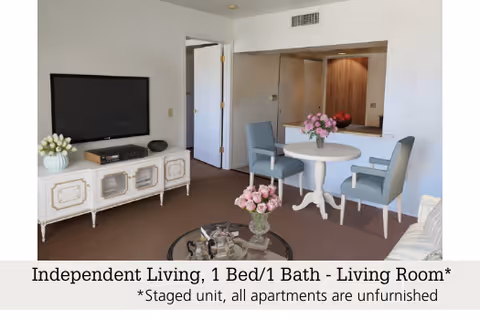 A staged living room in an independent living apartment featuring a white TV stand with a flat-screen TV, a round white table with two blue chairs, a glass coffee table with a flower arrangement, and a beige sofa partially visible. The room has white walls and brown carpet flooring.