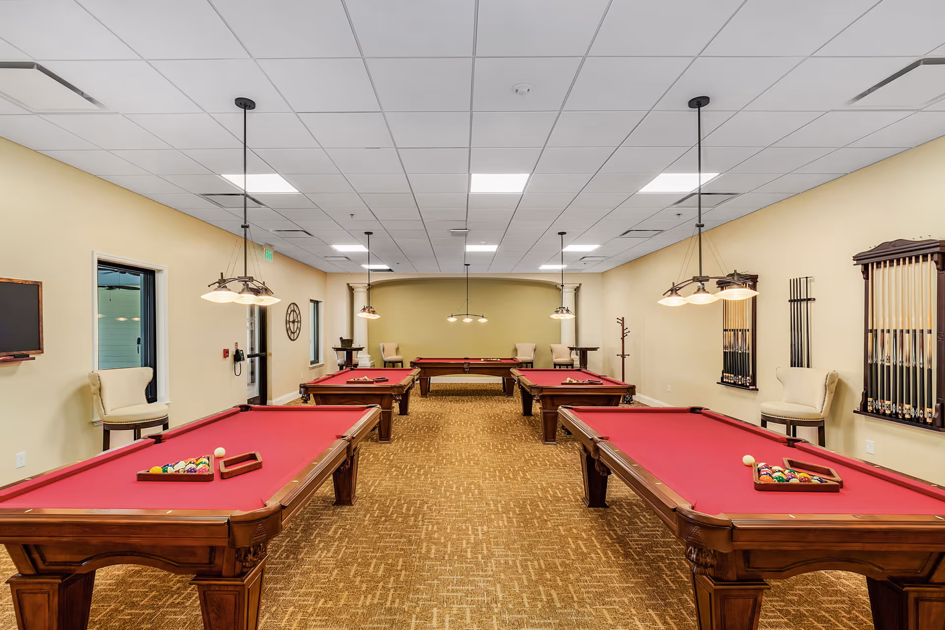 Interior view of a game room with four red felt pool tables arranged in two rows. The room has beige walls, carpeted floor, and ceiling lights hanging above each pool table. There are chairs and pool cue racks mounted on the walls.