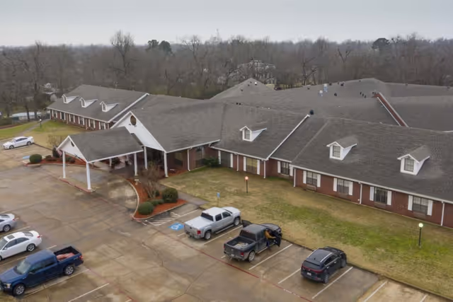Aerial view of a single-story brick building with a large covered entrance and multiple dormer windows on the roof. Several cars are parked in the parking lot in front of the building, which is surrounded by leafless trees and a grassy area.