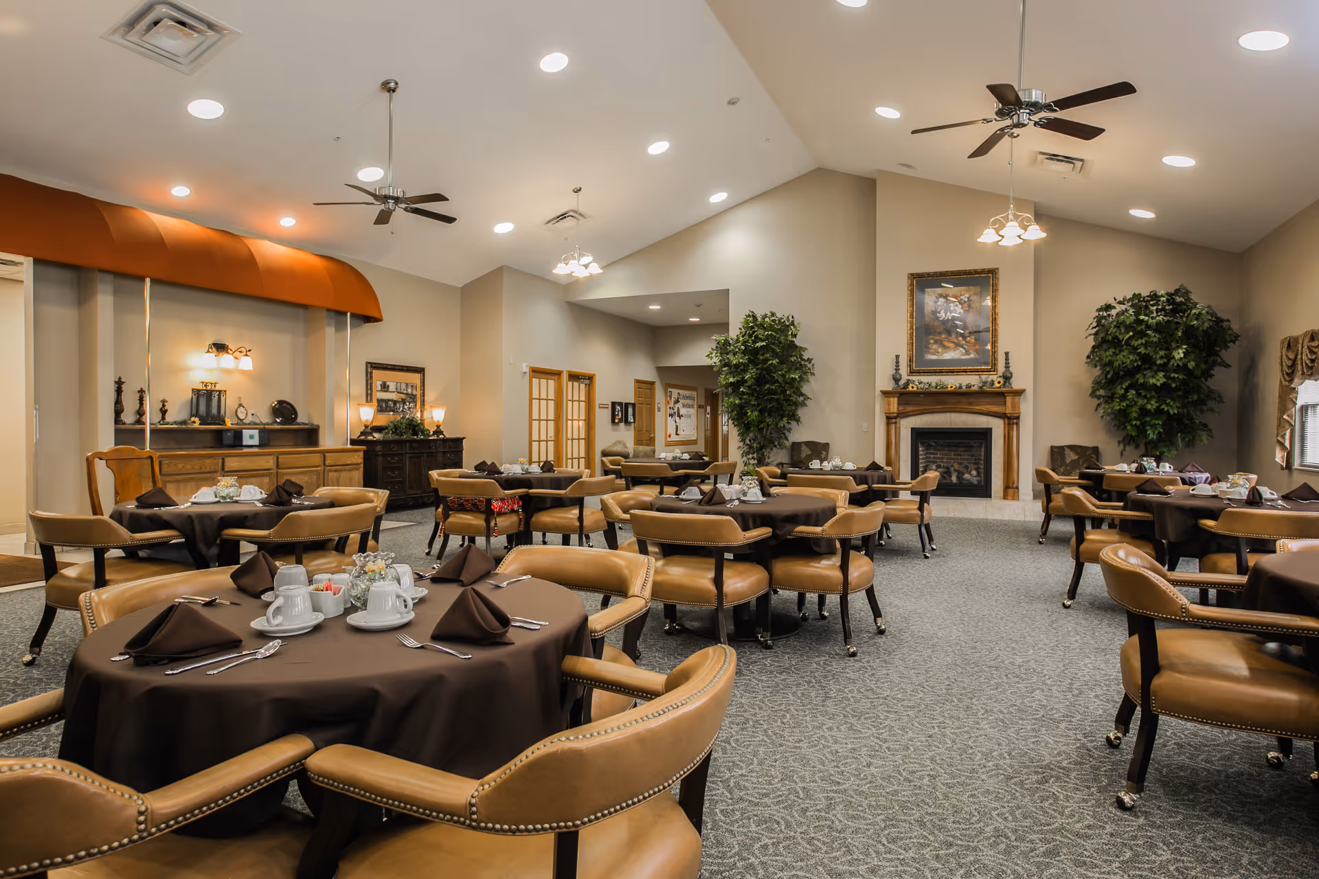 A spacious dining room with multiple round tables covered in dark brown tablecloths, each set with white cups, saucers, and folded napkins. The room features beige walls, a fireplace with a framed painting above it, ceiling fans, and large potted plants in the corners. The carpet is patterned in a neutral tone, and there is soft, warm lighting throughout.