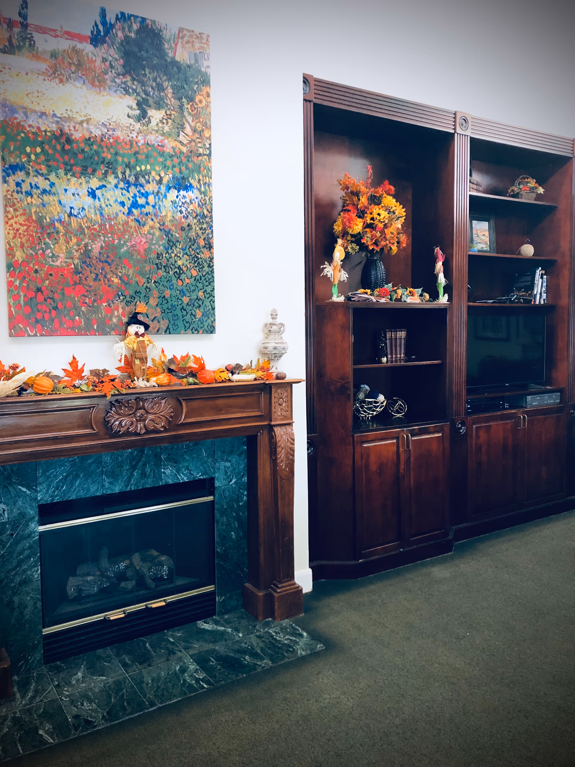 Cozy interior lounge with a decorated wooden fireplace mantel topped with autumn decorations and a large colorful painting, next to dark wood built-in shelves and a television.