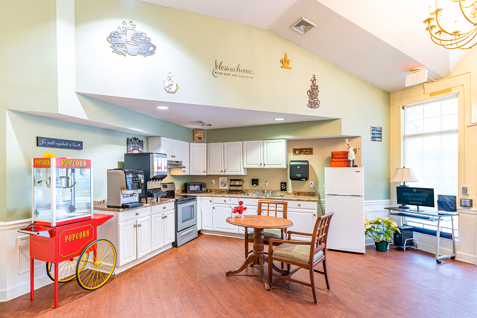 A bright and clean assisted living facility kitchen area with white cabinets, a stove, microwave, refrigerator, and a popcorn machine. There is a small round wooden table with two chairs in the center. The walls are decorated with coffee-themed wall art and a sign that reads 'bless our home with love and laughter.' A computer desk with a monitor displaying 'Brentwood at Elkhart Assisted Living' is near a large window letting in natural light.