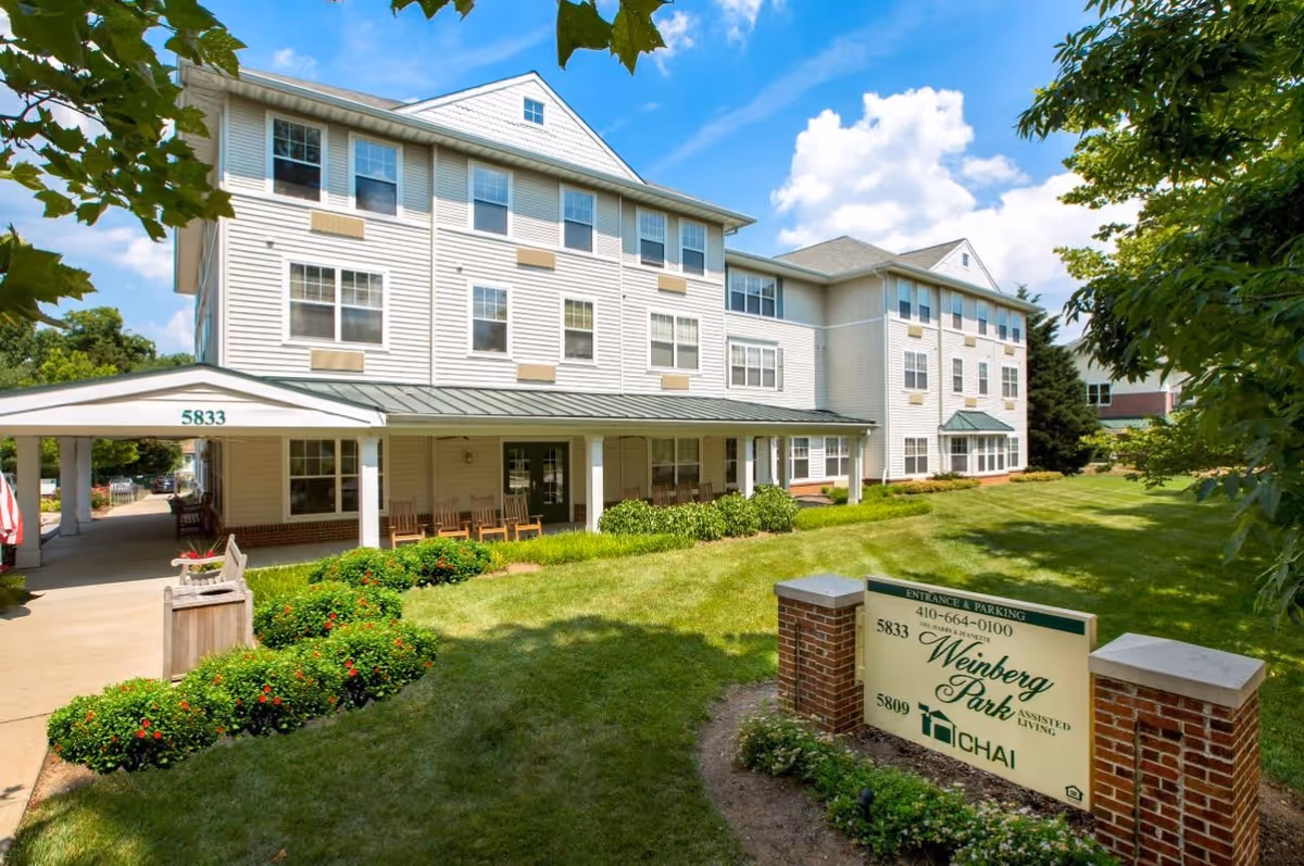 Exterior view of a three-story assisted living facility building with white siding and multiple windows. There is a covered entrance with rocking chairs and a well-maintained lawn with bushes and trees. A sign in front reads 'Weinberg Park Assisted Living' with contact information and the CHAI logo.