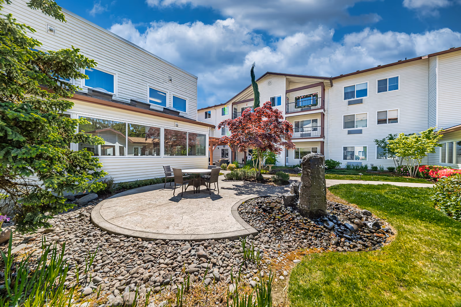 Outdoor courtyard area of a senior living facility with a round concrete patio featuring a table and four chairs, surrounded by landscaping including a small water fountain with rocks, green grass, and various trees and shrubs. The building has white siding and multiple windows under a partly cloudy blue sky.