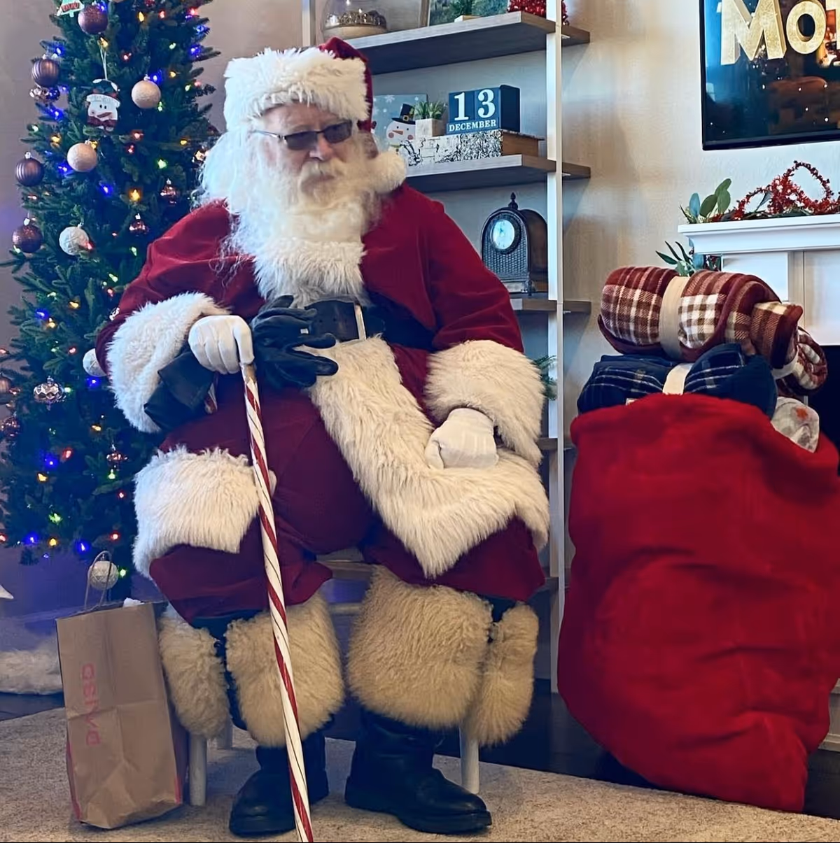 A person dressed as Santa Claus sits indoors by a decorated Christmas tree holding a candy-cane-striped staff beside a large red sack of gifts.
