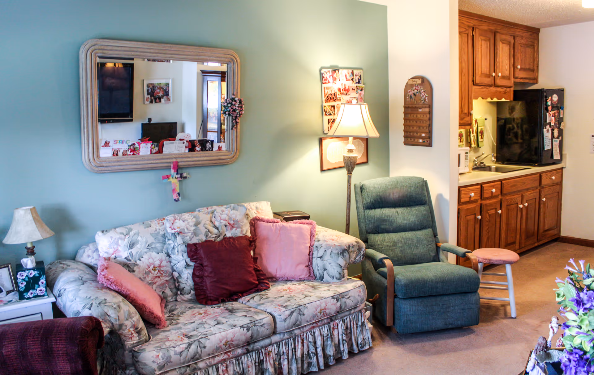 A cozy living room area with a floral patterned sofa adorned with pink and burgundy pillows, a green recliner chair, and a small wooden stool. The wall behind the sofa is painted light green and features a large mirror, a cross, a lamp, and a photo collage. To the right, there is a kitchenette with wooden cabinets, a black refrigerator, a microwave, and a sink.