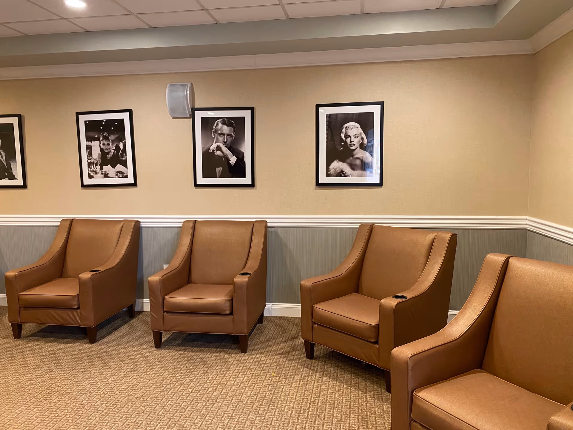 A row of brown leather armchairs in a seating area beneath framed black-and-white portraits on a beige wall.
