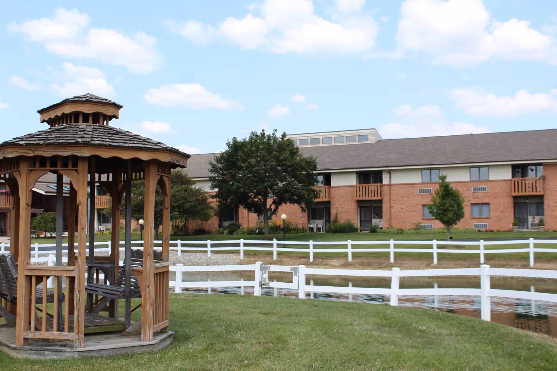 A wooden gazebo with benches inside sits on a grassy area near a small pond surrounded by a white fence. In the background, there is a two-story brick building with balconies and several trees under a partly cloudy sky.