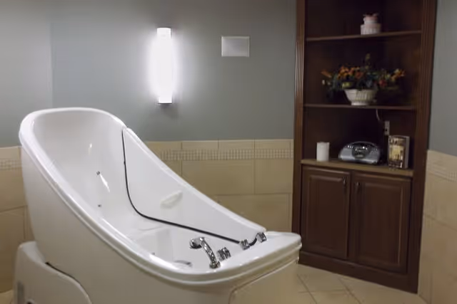 A modern, white therapeutic bathtub with chrome fixtures in a tiled bathroom. The walls are painted gray with beige tiles halfway up, and there is a wooden cabinet with shelves holding decorative items and a radio on the right side. A wall-mounted light fixture is above the bathtub.
