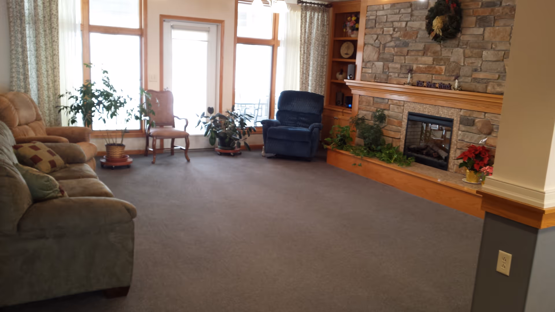 A cozy living room with a stone fireplace decorated with a wreath and plants. The room has a green sofa, a blue recliner, a wooden chair, and several potted plants near large windows with curtains. There is a built-in wooden shelf next to the fireplace.