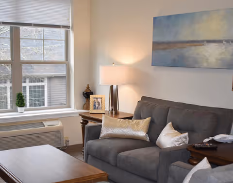 Cozy living room with a gray loveseat topped with decorative pillows, side tables with a lamp and framed photo, a large window, and wall art.