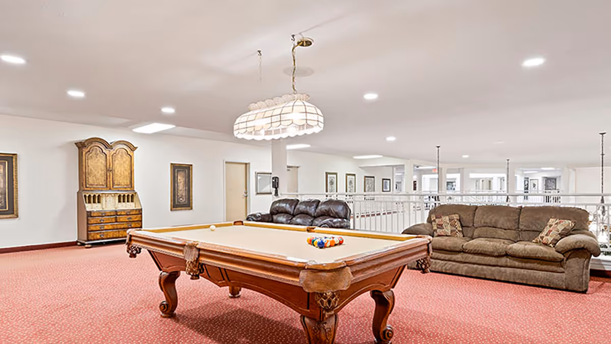 A spacious recreational room with a pool table in the center, a brown leather couch and a brown fabric couch with patterned pillows along the wall, a wooden cabinet, framed artwork on white walls, and a large overhead light fixture above the pool table. The room has red carpet flooring and bright ceiling lights.