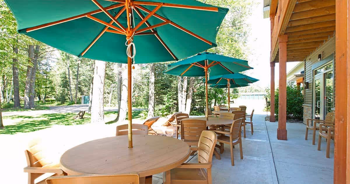 Outdoor patio area at Milestone Senior Living Eagle River with round wooden tables and chairs under large green umbrellas, surrounded by trees and greenery.