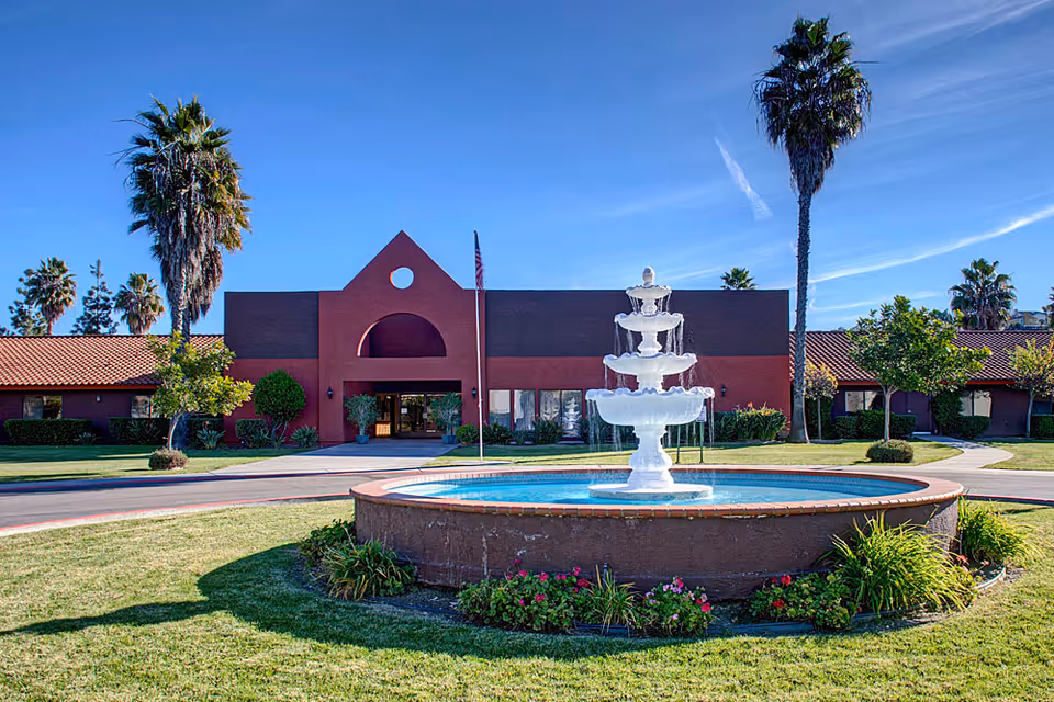 Front exterior view of Santa Fe Post Acute facility with a large circular fountain in the foreground, surrounded by grass and flowers. The building has a red and brown facade with a tiled roof, palm trees, and a clear blue sky above.