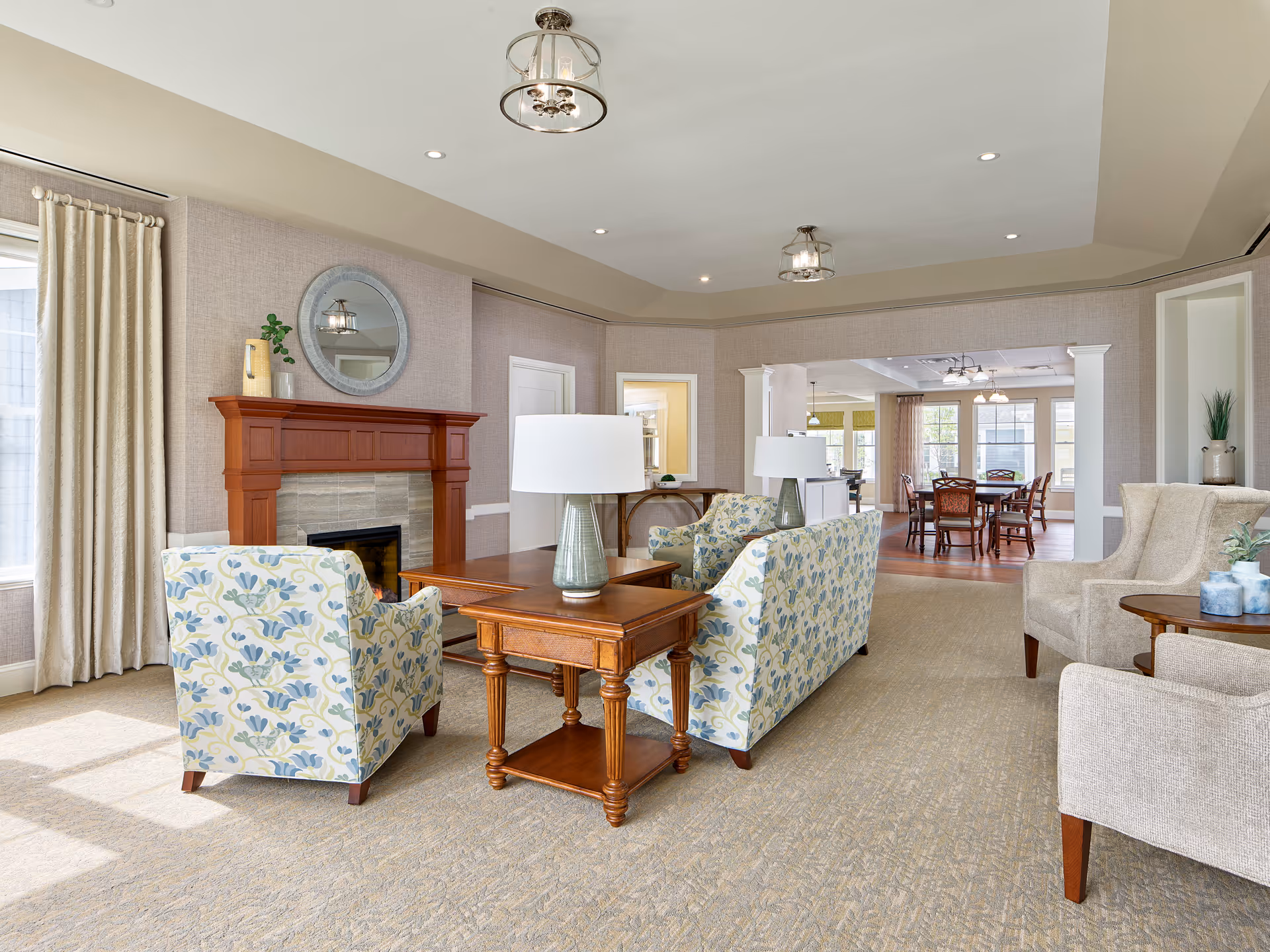 A bright and spacious living room area in a retirement community featuring a fireplace with a wooden mantle, a round mirror above it, two floral patterned armchairs, a wooden coffee table with a lamp, beige carpet, and large windows with curtains allowing natural light to fill the room. In the background, there is a dining area with a wooden dining table and chairs.