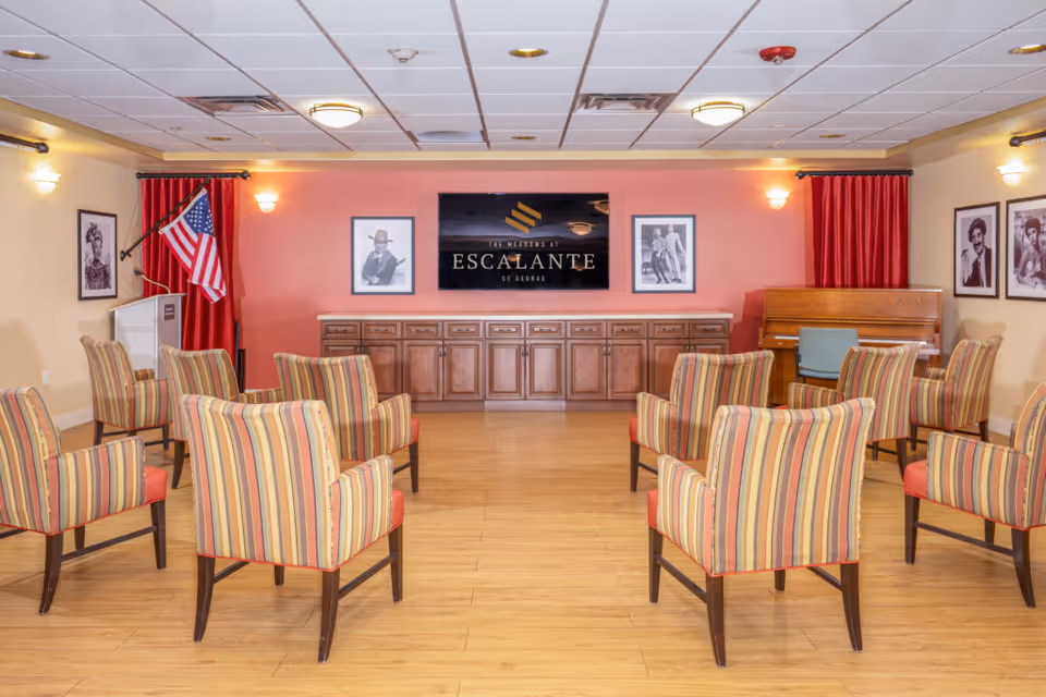 Activity room with striped armchairs arranged facing a wall with a sign that reads 'Escalante', framed photos, a piano and an American flag.