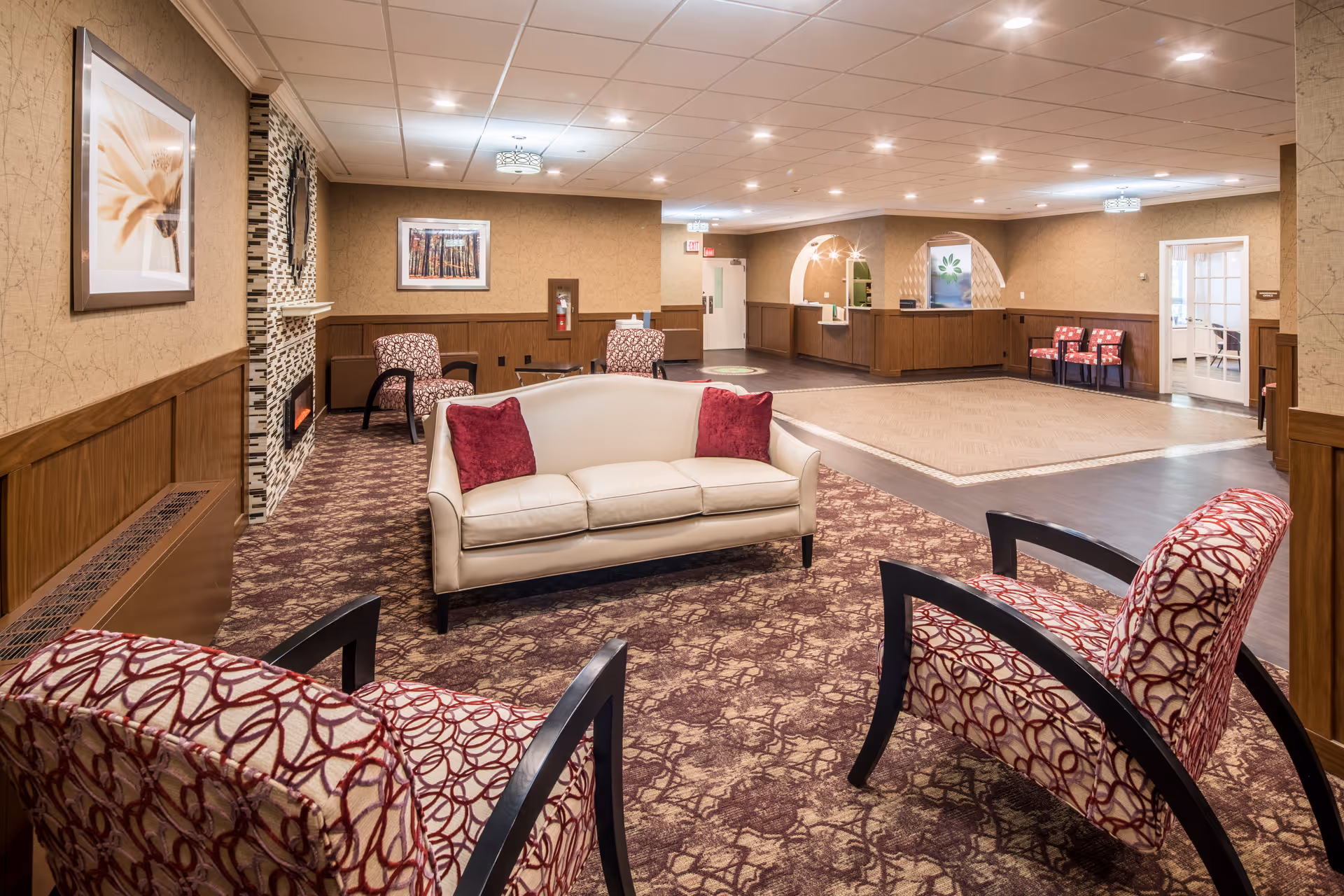 A spacious and well-lit living room area in a senior living facility featuring a white sofa with two red pillows, two patterned armchairs, a fireplace with a stone surround, framed artwork on the walls, and a reception desk in the background.