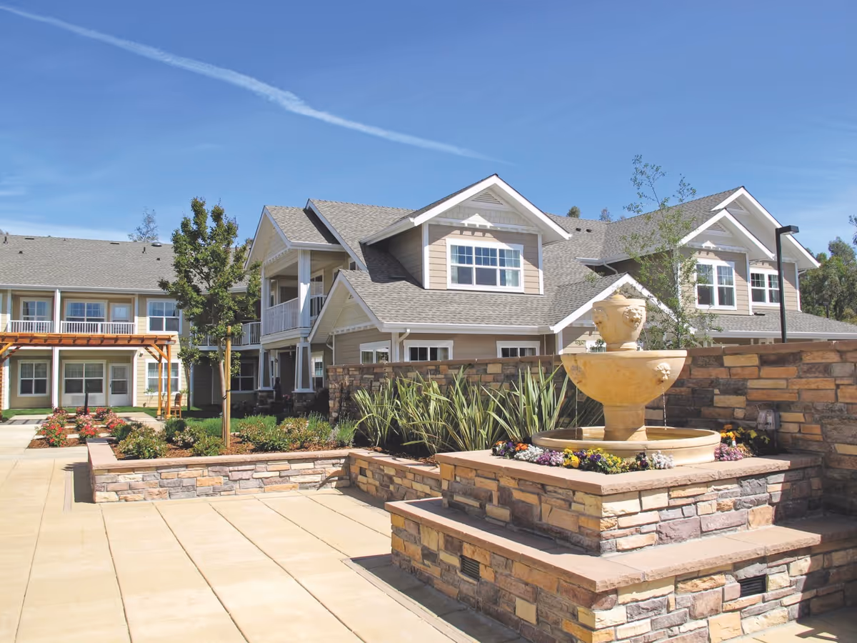 Outdoor courtyard area of Camden Springs Gracious Retirement Living featuring a stone water fountain surrounded by flowers, paved walkways, landscaped garden beds with plants and trees, and beige two-story residential buildings with white trim under a clear blue sky.