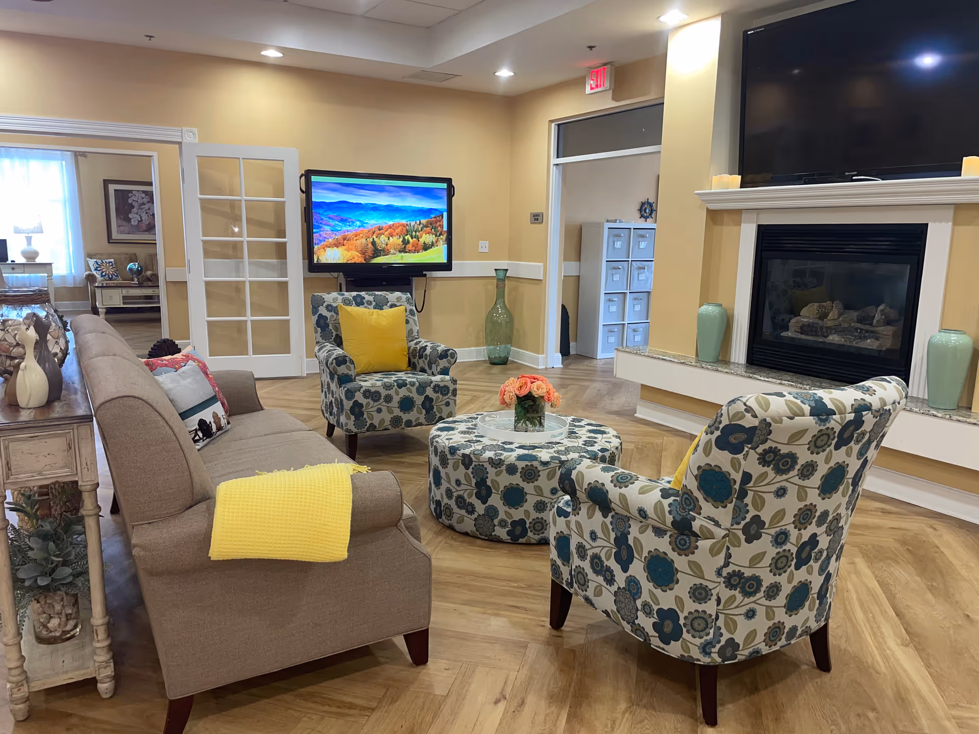 A cozy living room with a beige sofa adorned with colorful pillows and a yellow throw blanket, two floral-patterned armchairs with yellow cushions, and a matching round ottoman with a vase of flowers on top. The room features a fireplace with a large TV mounted above it, light wood flooring, and soft yellow walls. French doors lead to another room with additional seating and decor.