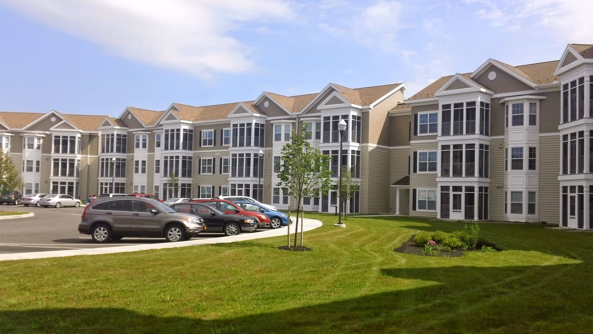 Three-story senior living apartment building with many windows, a curved driveway, parked cars, and a grassy lawn in front.