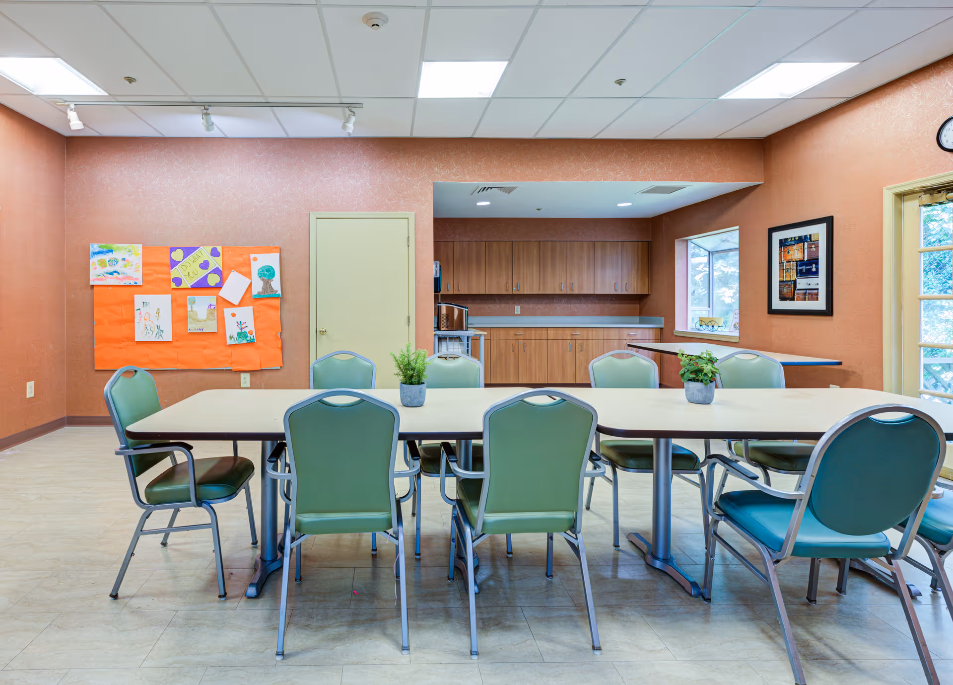 A communal room with a long table surrounded by green chairs. The walls are painted a warm brown color, and there is a bulletin board with colorful drawings on the left wall. In the background, there is a kitchenette area with wooden cabinets and a countertop. Two small potted plants are placed on the table. A window and a door letting in natural light are visible on the right side.