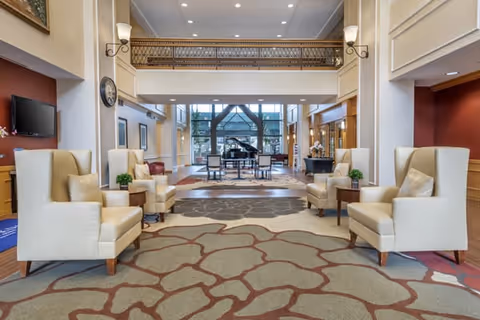 Spacious two-story lobby with pairs of armchairs, a grand piano, balcony railing and large windows.