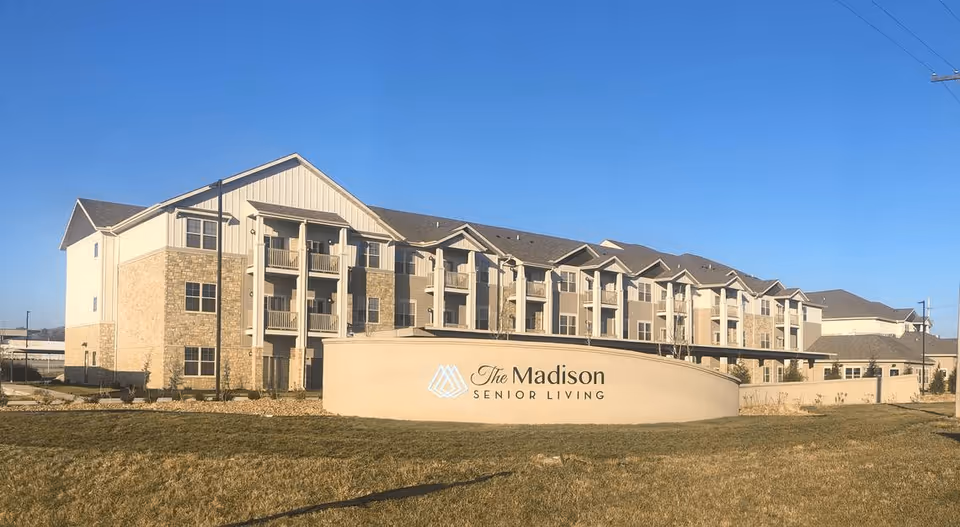 Exterior view of a three-story senior living building with balconies and a front sign reading "The Madison Senior Living".
