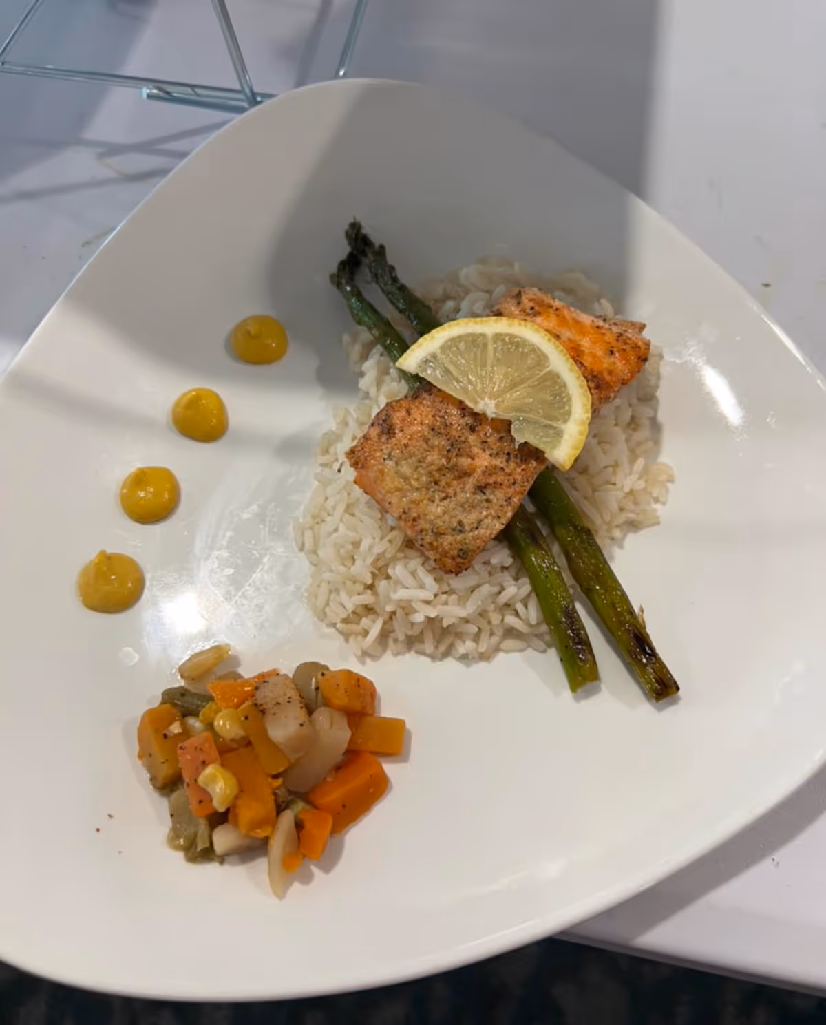Plated seared salmon with a lemon slice on rice and asparagus, served with mixed vegetables and mustard dots on a white plate.