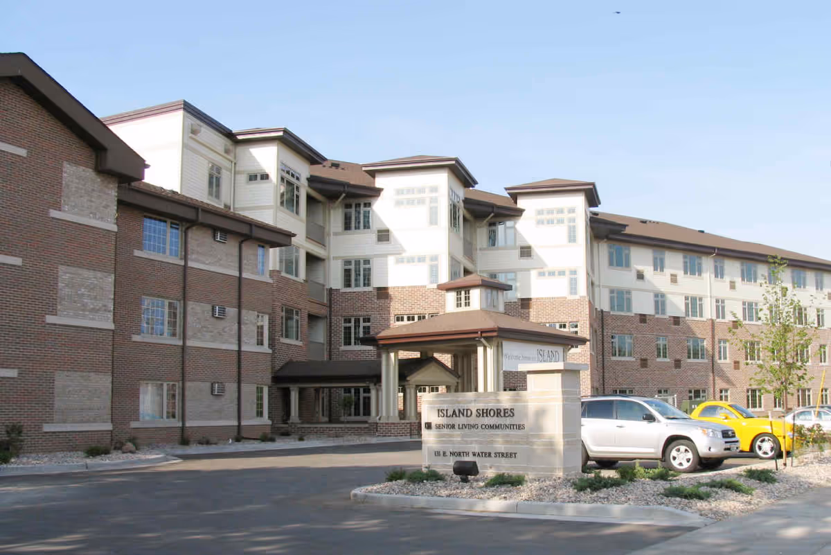 Exterior view of a multi-story senior living community building named Island Shores with a parking area in front and a sign displaying the facility name and address.