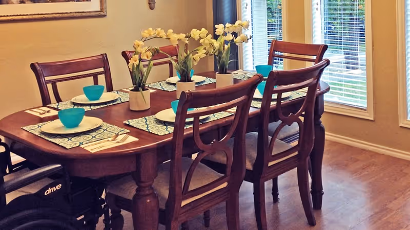 A wooden dining table set for four with blue bowls, white plates, and floral placemats. Three small potted plants with yellow flowers are placed in the center of the table. The room has wooden flooring and large windows with white blinds, allowing natural light to enter. A wheelchair is partially visible next to the table.