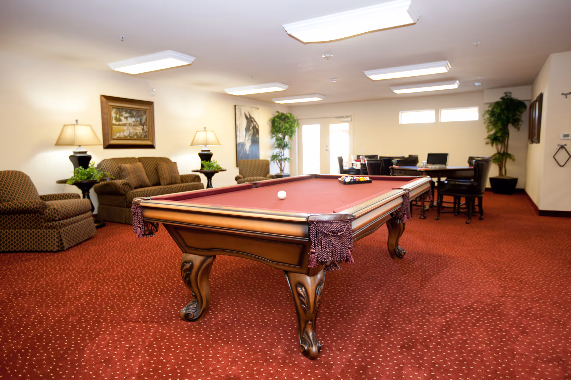 A spacious room with a red carpet featuring a pool table with a red felt surface in the center. The room has comfortable seating including armchairs and a sofa, side tables with lamps and plants, a dining table with chairs in the background, framed artwork on the walls, and potted plants near the windows and door.
