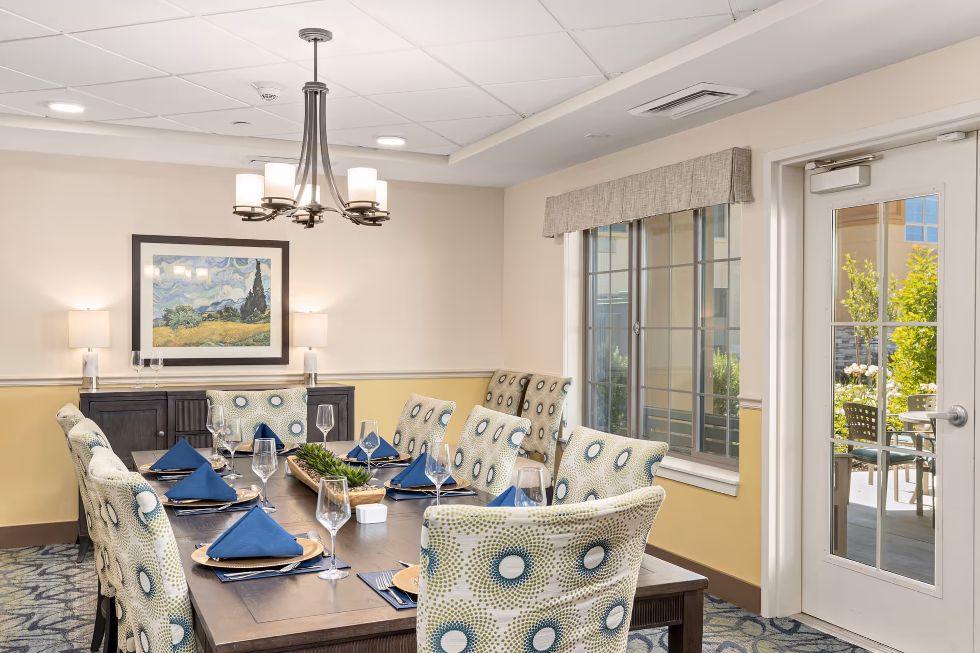 A well-lit dining room with a rectangular wooden table set for eight people. The chairs have patterned upholstery with circular designs in blue and green. Each place setting includes a gold charger plate, blue napkin, and clear wine glass. A modern chandelier hangs above the table. On the wall is a framed painting and two table lamps on a dark wooden sideboard. A window and glass door reveal an outdoor patio area with chairs and greenery.