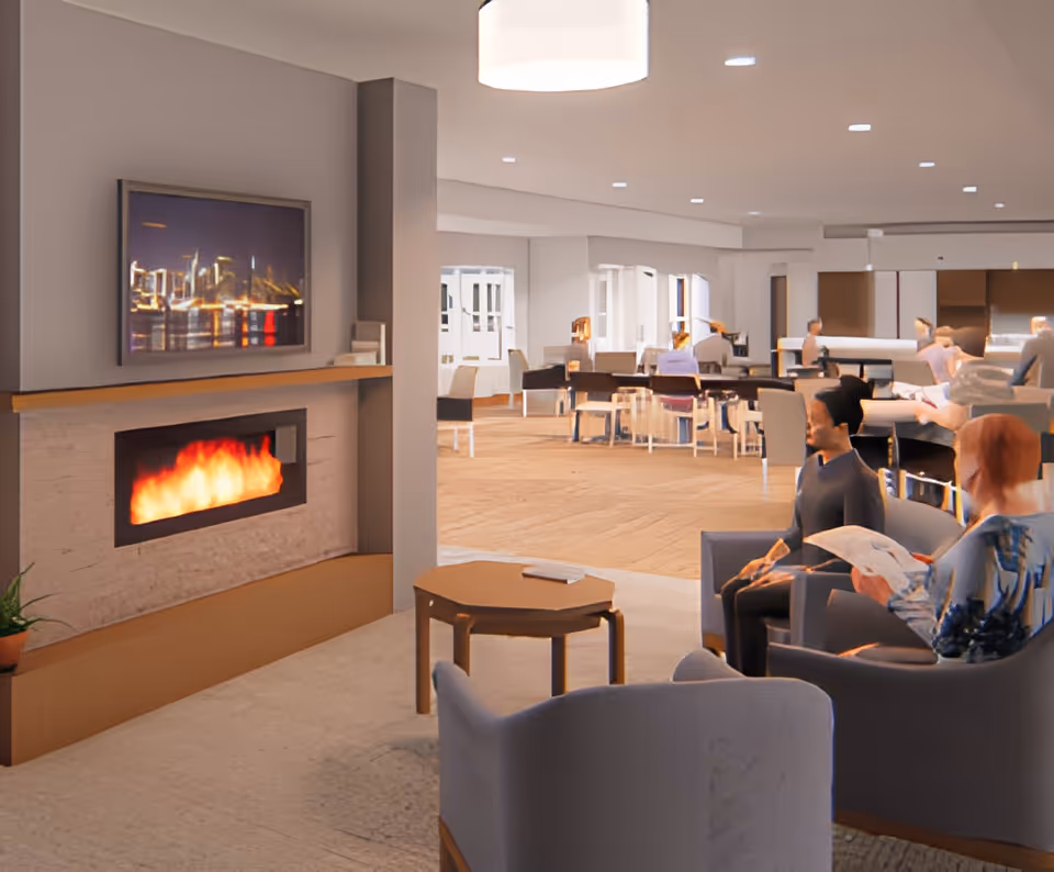 A cozy senior living common area with a modern fireplace on the left wall, a framed cityscape picture above it, and several seniors seated in comfortable chairs. The background shows a spacious dining area with tables and chairs, and more seniors engaged in activities.