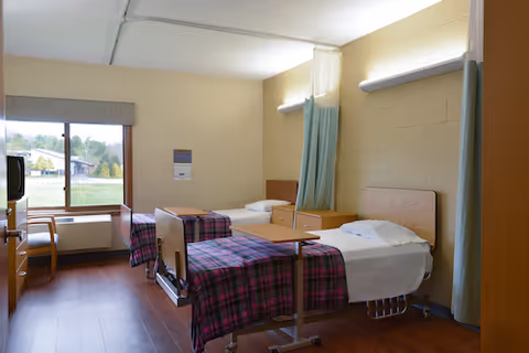 A nursing home bedroom with two hospital-style beds with plaid blankets, bedside tables, a window, and seating.