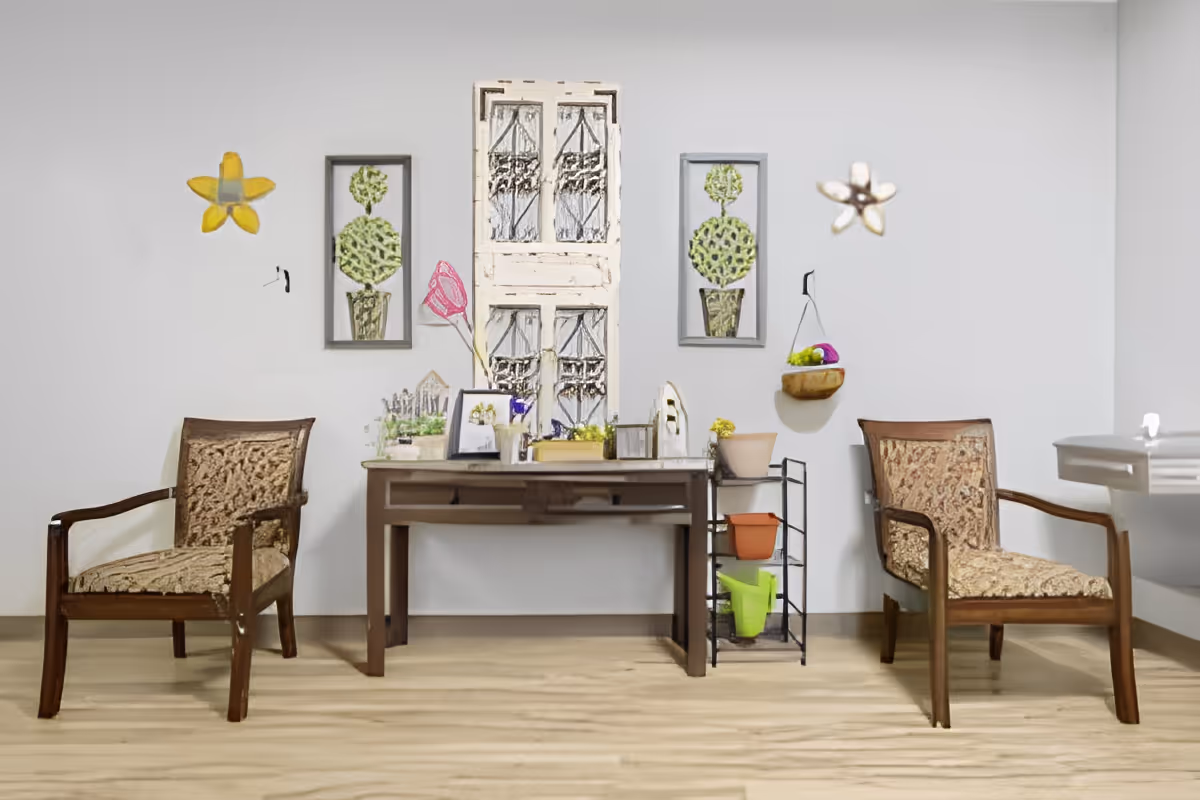 A cozy interior space featuring two wooden armchairs with patterned cushions on either side of a wooden table. The table holds various decorative items including framed pictures, small plants, and a miniature house model. On the wall behind the table are two framed topiary artworks and a decorative window panel. The floor is light wood, and there is a small black metal shelf with colorful pots next to the table.