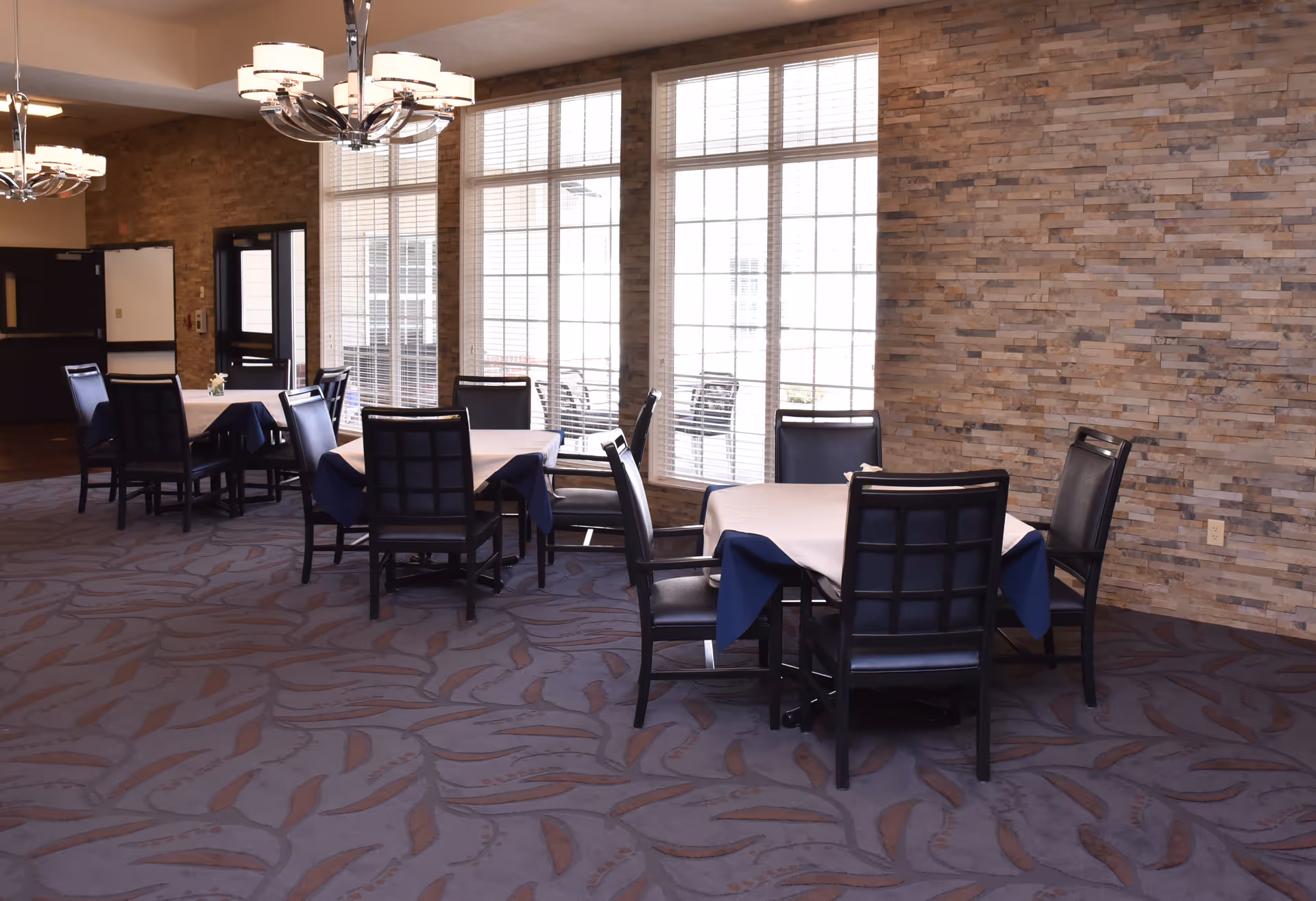 A dining room with tables draped in white and blue tablecloths, black chairs, large windows, and a stone accent wall.