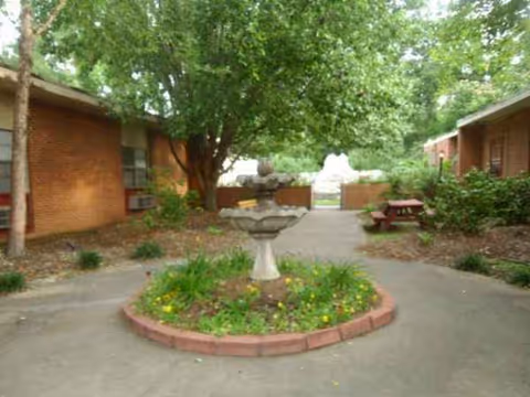 Outdoor courtyard with a stone fountain, flower bed, trees, and seating between brick buildings.