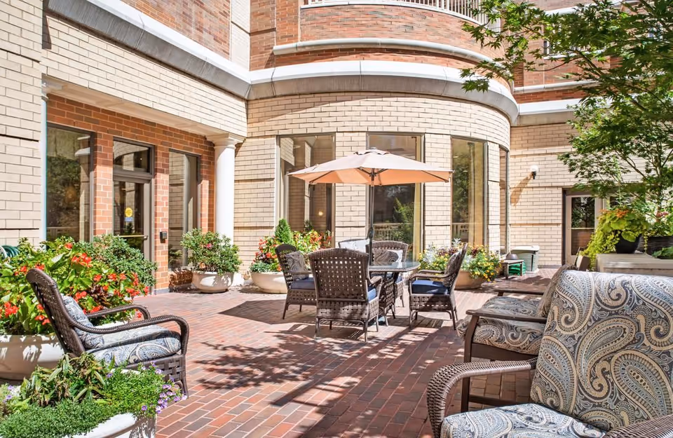 Sunlit brick courtyard with wicker patio chairs and a table under an umbrella, surrounded by potted plants in front of a curved brick building.