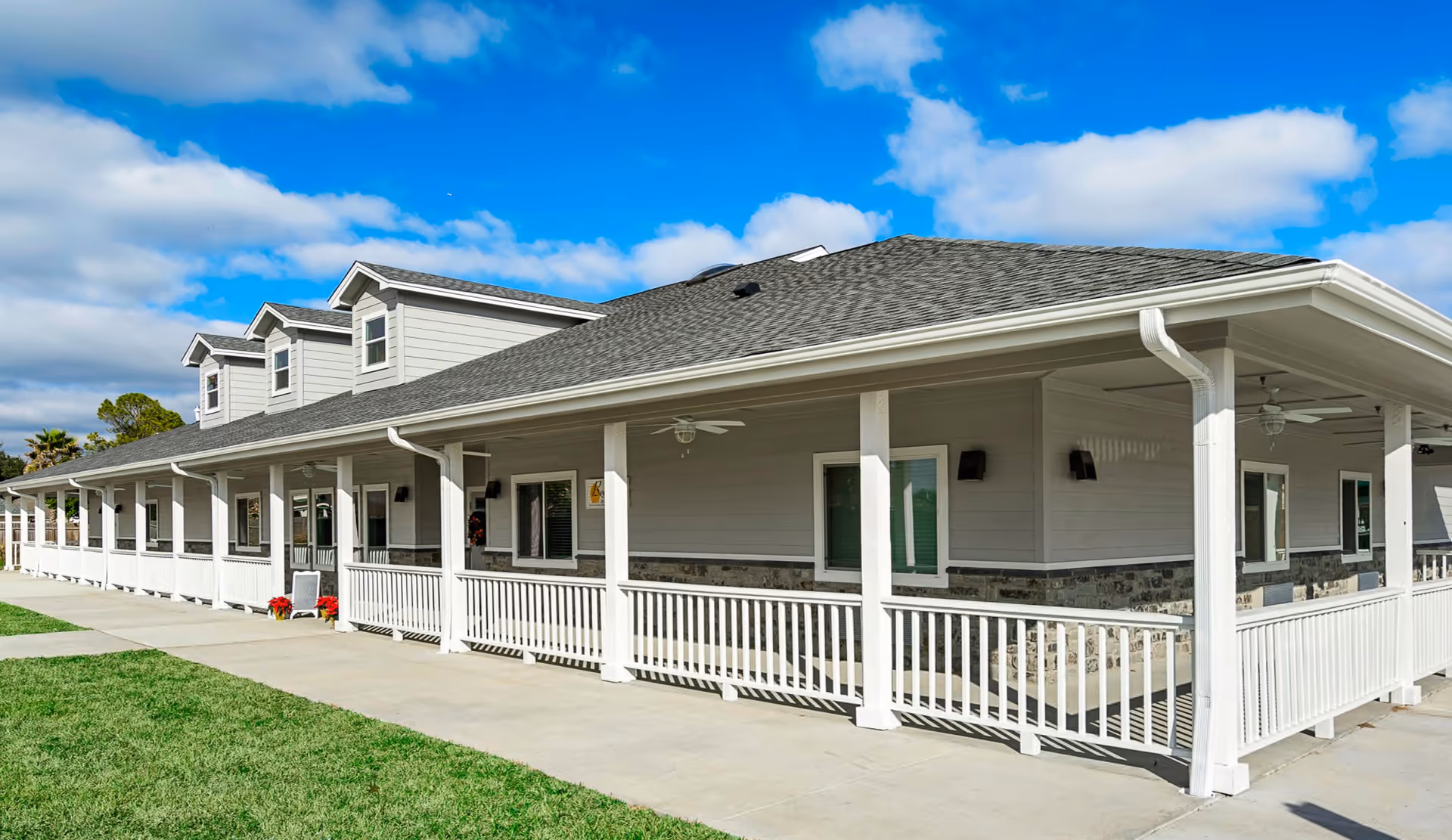 Exterior view of a single-story building with a gray roof, light gray siding, and white railings along a covered porch. The building has several windows and dormer windows on the roof. The sky is blue with some clouds, and there is a green lawn in the foreground.