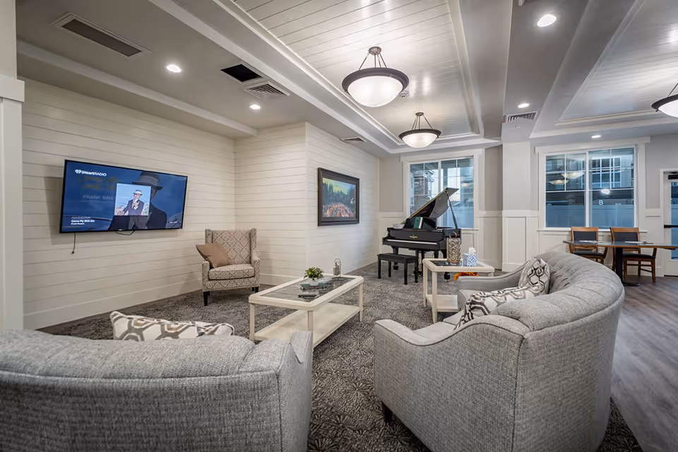 A cozy senior living common area with two curved gray sofas facing a wall-mounted TV displaying a Frank Sinatra image. There is a patterned armchair, two white coffee tables with decorative items, a grand piano near the windows, and a small dining table with chairs in the background. The room has white paneled walls, recessed lighting, and large windows letting in natural light.