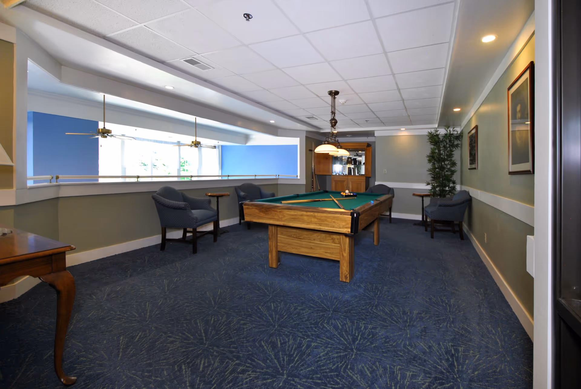 Interior room with a pool table in the center, surrounded by four blue upholstered chairs and small wooden side tables. The room has blue patterned carpet, light green walls with framed pictures, ceiling lights, and a large window with ceiling fans visible through it.