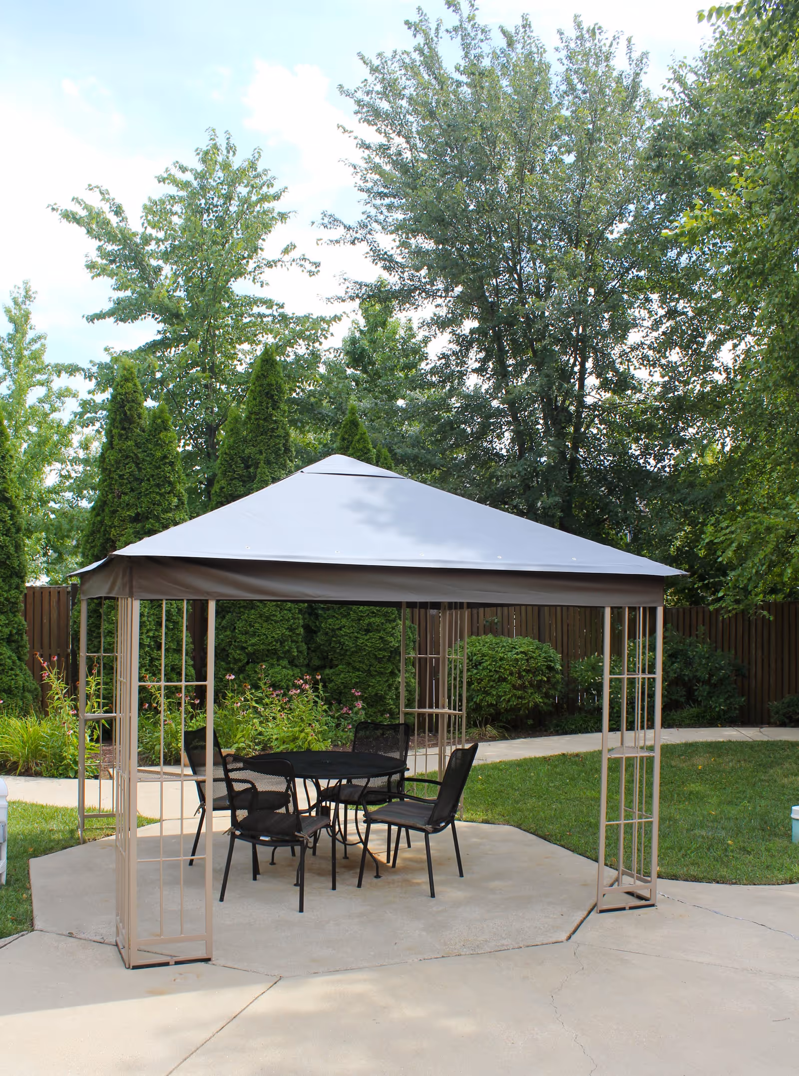 Outdoor patio area with a beige gazebo covering a black metal table and four matching chairs, surrounded by green grass, bushes, and tall trees in the background.