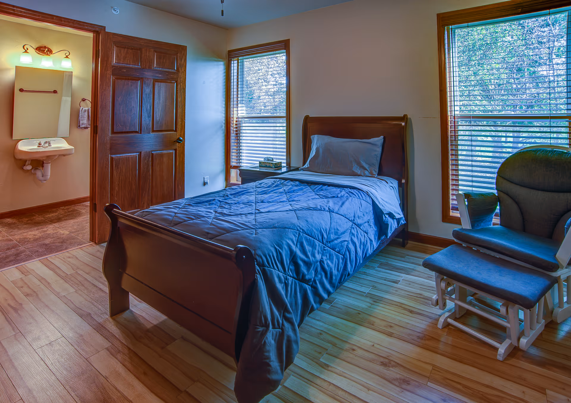 A small bedroom with a single wooden bed made with a blue comforter and pillow. There are two windows with wooden blinds letting in natural light. To the right of the bed is a cushioned rocking chair with an ottoman. The room has wooden flooring and beige walls. An open door reveals a small bathroom area with a sink, mirror, and towel rack.