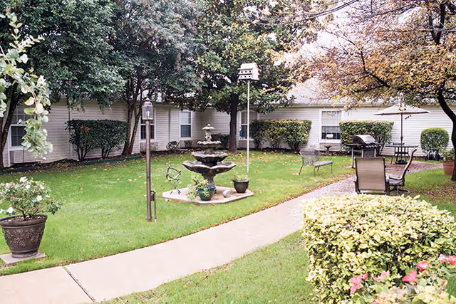 Outdoor courtyard area with green grass, trees, shrubs, and a concrete walkway. There is a multi-tiered water fountain in the center, a birdhouse on a pole, metal benches, a table with chairs, and potted plants. The building exterior with white siding and windows surrounds the courtyard.