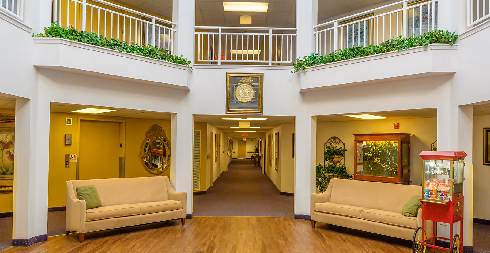 Atrium-style common area with two sofas, plants, balcony railings and a central hallway in a senior living facility.