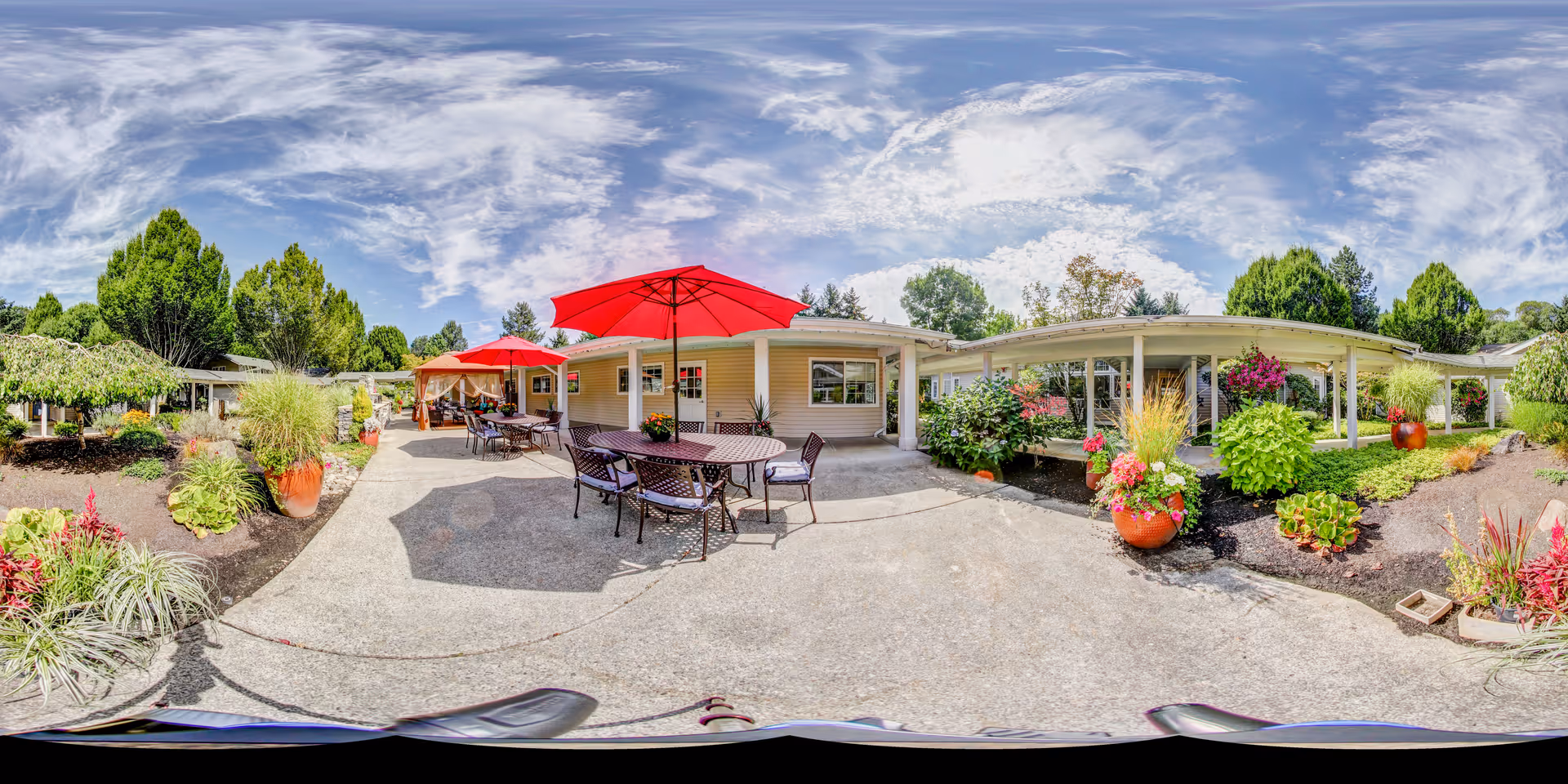 Outdoor patio area at Aegis Living Issaquah featuring several tables with red umbrellas, surrounded by landscaped garden beds with various plants and flowers under a partly cloudy sky.