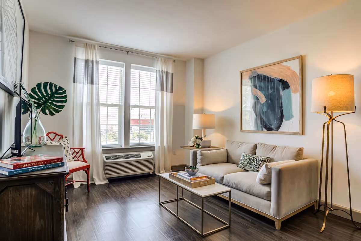 A bright living room with a beige sofa adorned with patterned pillows, a modern floor lamp, and a side table with a lamp and flowers. A large abstract painting hangs on the wall above the sofa. In front of the sofa is a rectangular coffee table with books and a small plant. To the left, there is a wooden cabinet with books and a glass vase with a large green leaf, and a red chair near a window with white and gray curtains. The floor is dark wood, and sunlight streams through the window.