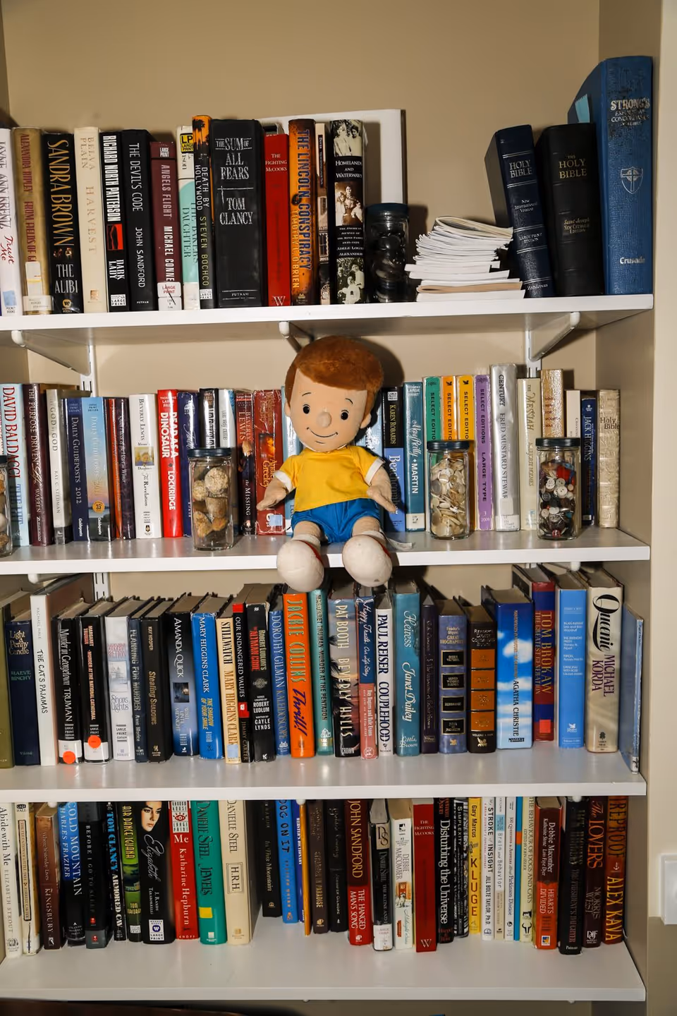 White bookshelves packed with books, jars of shells and buttons, and a small plush doll seated on the middle shelf.